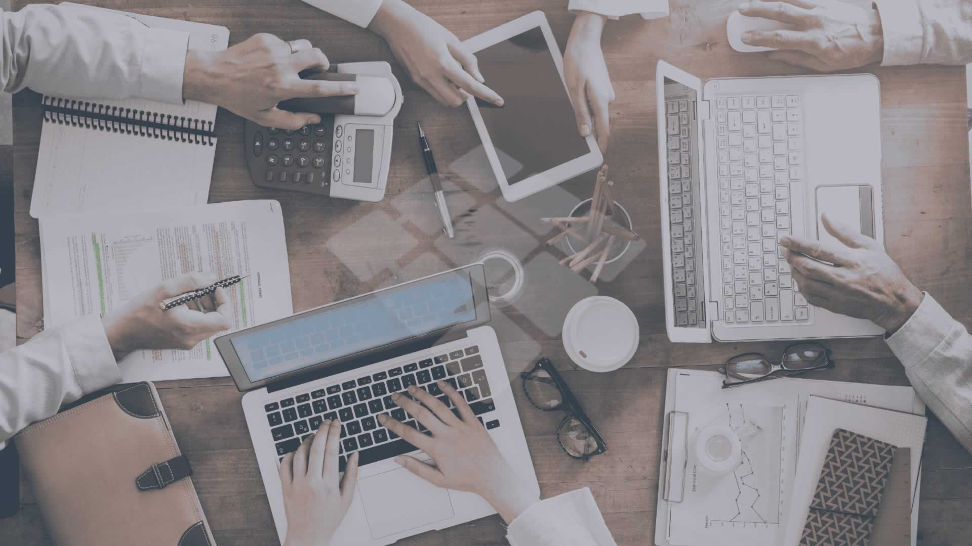 Mesa de trabalho com várias pessoas reunidas, notebooks abertos, telefones fixos, canetas, agendas e óculos dispostos sobre a superfície